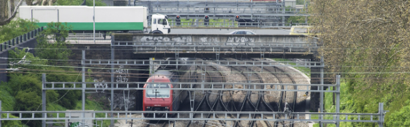 Voies et train qui entre en gare en dessous d'un pont où circulent des voitures et des camions.